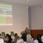 Karin Fernschlld hält das Mikro in der Hand und spricht zum Publikum. Groß links im Bild die Projektion des Veranstaltungsplakats.